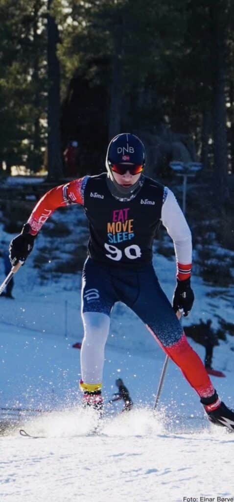 Cross-country skiing athlete racing on snow wearing FF Skis gear and ‘Eat Move Sleep’ race bib, forest background.