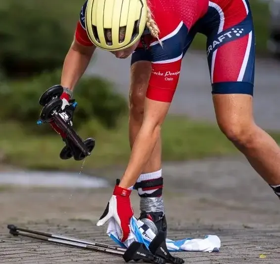 Victoria-nitteberg-ff-sprint-med norsk treningsdrakt med rulleski fra FFskis i hånden plukker opp et par staver fra asfalten.